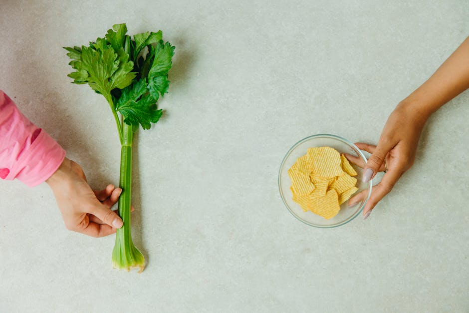 A top-down view of hands holding celery and chips on a light background, emphasizing a healthy versus unhealthy snack choice.