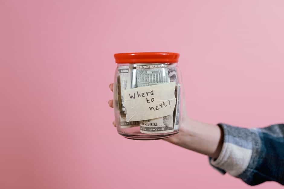 A hand holding a glass jar filled with dollar bills labeled 'Where to next?' against a pink background, symbolizing savings and future planning.