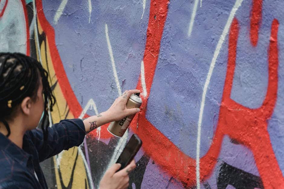 From above side view of crop faceless female artist with braided hair painting graffiti on wall while standing on street