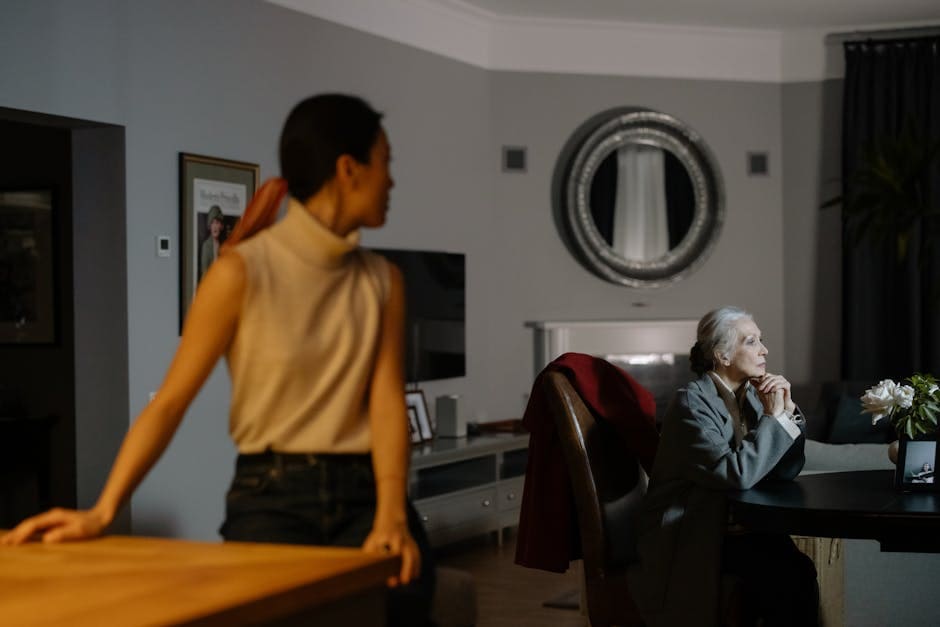 Senior woman deep in thought at a table with a younger woman in focus background, evoking a quiet, introspective mood.