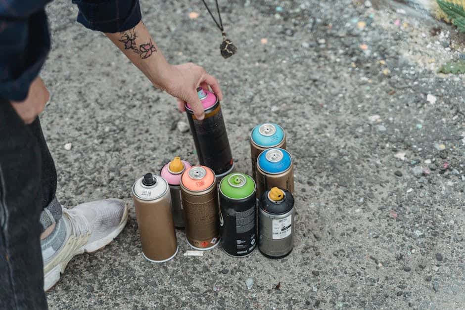 Crop anonymous person in sneakers with tattoo and heap of multicolored spray paint cans on ground standing on street in city