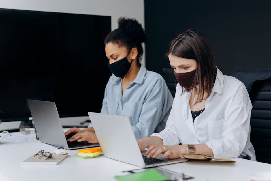 Two women wearing face masks working on laptops in a modern office environment.